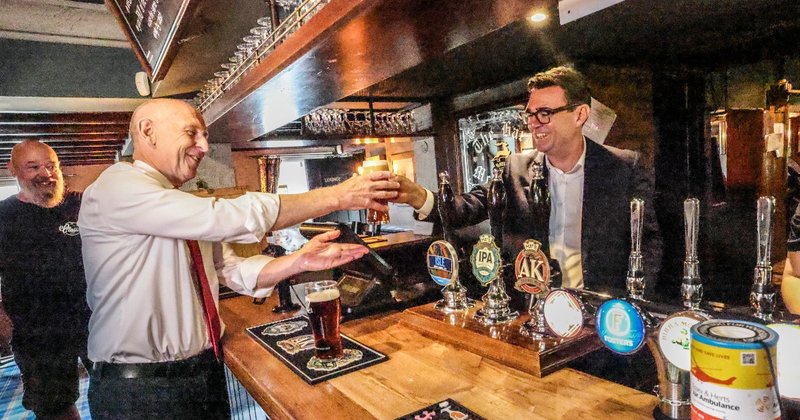 Prime Minister Andy Burnham handing Chancellor John Healey a pint during a visit to a pub.