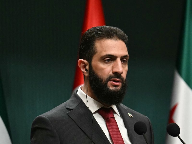 A photo of Syria’s President Ahmed al-Sharaa speaking at a podium with Syrian and Turkish flags in the background.