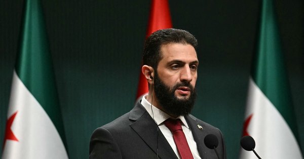 A photo of Syria’s President Ahmed al-Sharaa speaking at a podium with Syrian and Turkish flags in the background.