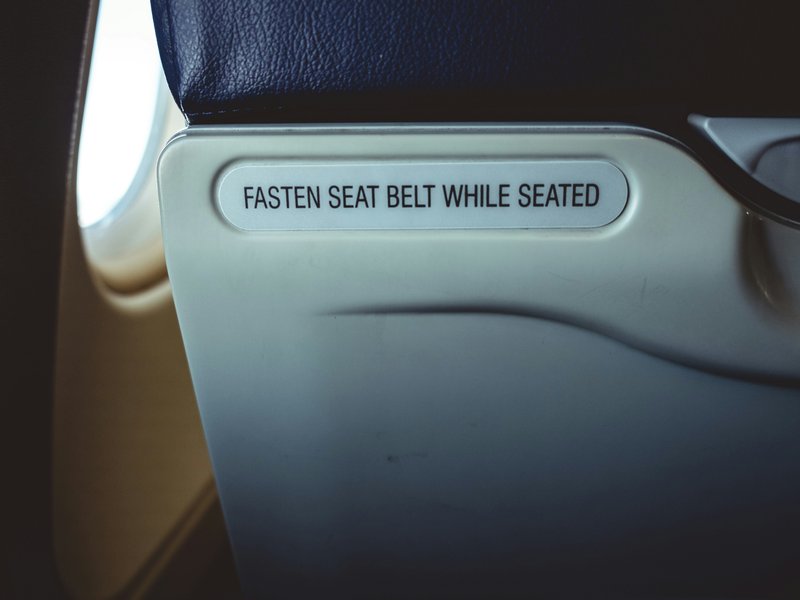 Back of a seat on an aeroplane with a sign that reads “fasten seat belt while seated”.