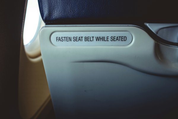 Back of a seat on an aeroplane with a sign that reads “fasten seat belt while seated”.