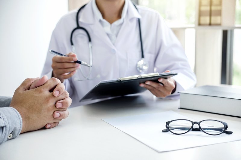 Doctor sits across a table from a patient, holding a clip board and pen.