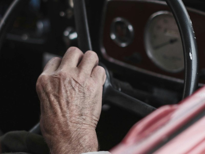 An older man’s hand on the steering wheel of a car.