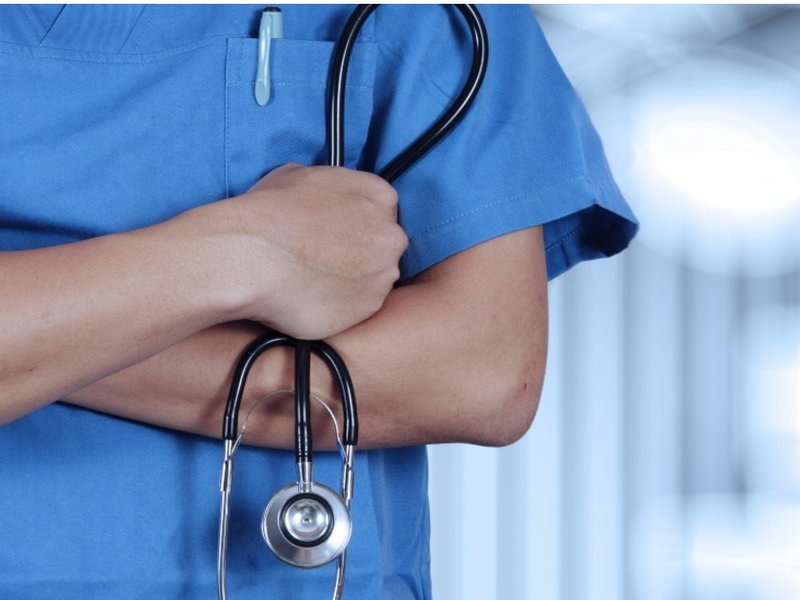 A person wearing blue medical scrubs with their arms folded and holding a stethoscope.